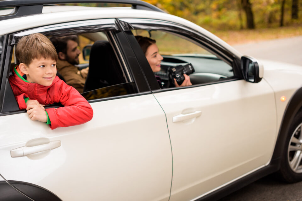 Cute smiling boy looking through car window while traveling with parents in autumn forest